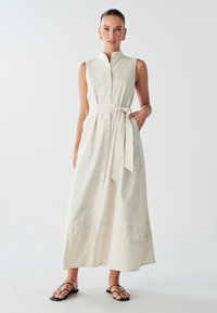 Svetlo béžové, bezrukávové maxi šaty s gombíkmi, so zúženým pásom a volánovou sukňou. Tkanina má textúrovaný povrch; kombinované s čiernymi sandálmi.