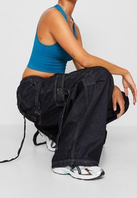 Dark denim cargo pants with multiple pockets, stitched accents, loose fit, paired with a teal ribbed crop top and white sneakers.