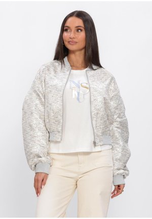 Vrouw met lang donker haar, gekleed in een zilverkleurig bomberjack met textuur, een wit T-shirt met grafisch ontwerp en beige broek, staand tegen een witte achtergrond.