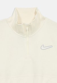 Krémový Nike pulóver s vysokým golierom, polovičným zipsom a šedou výšivkou loga; mäkká textúra a ľahký materiál.
