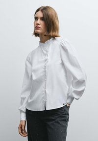 White blouse with a high collar, ruffled front, and long puffed sleeves. Crafted from a smooth fabric, features button closure and a curved hemline.
