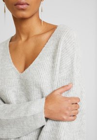 Hellegrauer gestrickter Pullover mit V-Ausschnitt, der eine gerippte Textur und lockere Ärmel zeigt. Verfügt über eine glatte Oberfläche und einen schichtenartigen Look.