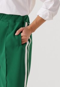 Groene sportbroek met een zijzak en witte strepen. De stof heeft een gladde textuur en de tailleband is elastisch.