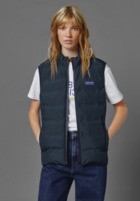 Marinblå puff väst med hög krage, med dragkedja framtill och logopatch, ihop med en vit t-shirt och blå jeans. Slät textur.