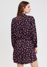 Vestido negro con flores y mangas largas, que presenta un patrón de flores en rosa y morado, cintura ceñida con cinturón y una falda ligeramente acampanada.