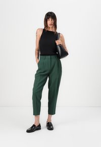 Green tailored trousers with a high waist and side pockets, paired with a black ribbed tank top and black loafers. Black handbag accent.