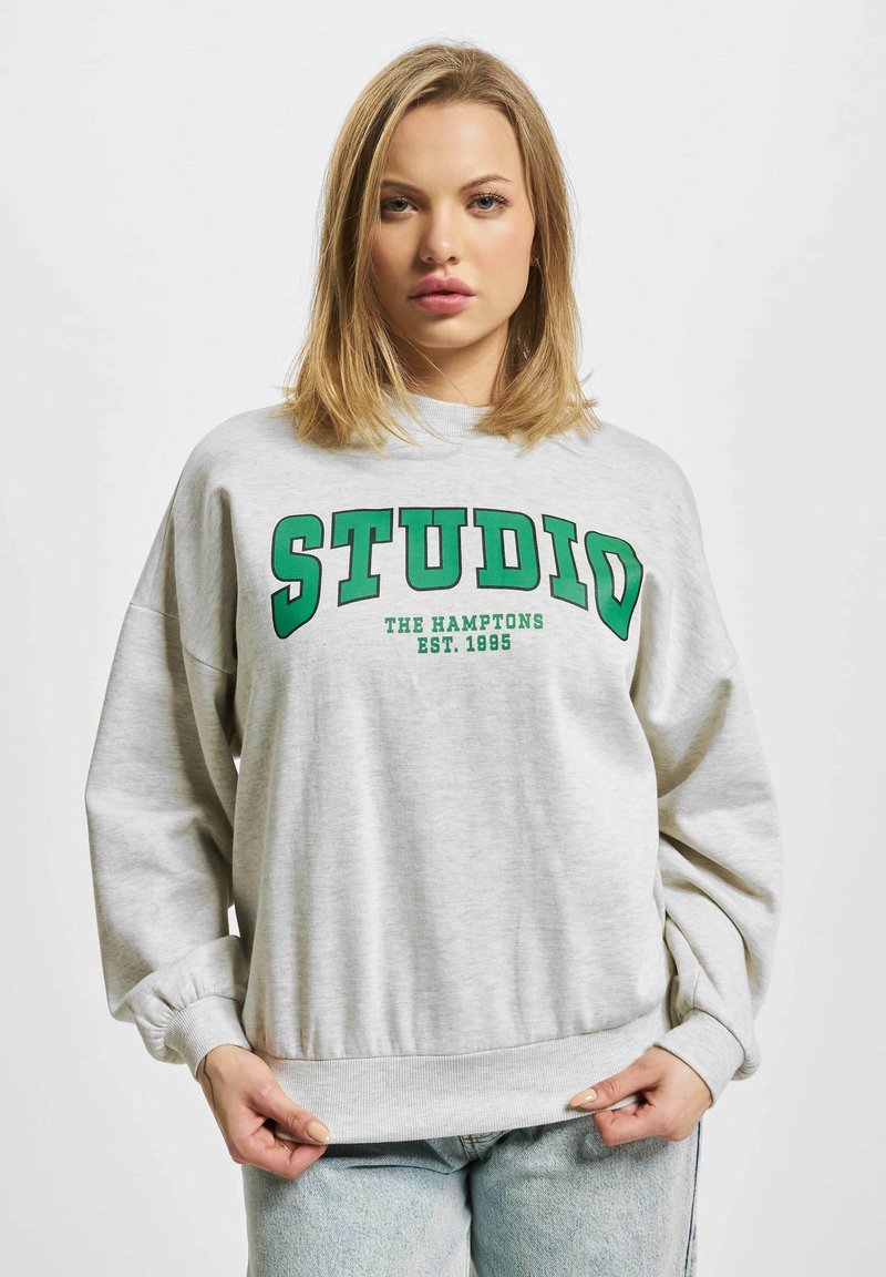 Grå hettegenser med grønn tekst "STUDIO THE HAMPTONS EST. 1995." Har en løs passform, ribbede mansjetter og et båndet hem.