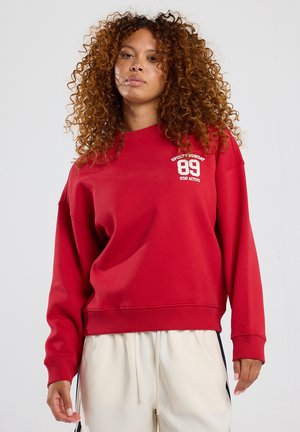 Punainen huppari, jossa on pyöreä pääntie ja ylisuuri malli. Vasemmassa rinnassa valkoista tekstiä ja numeroita. Pehmeää, teksturoitua kangasta.