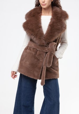 Donna che indossa un gilet in ecopelliccia marrone con cintura sopra un maglione bianco a coste e jeans blu a gamba larga, in piedi con una mano in tasca.