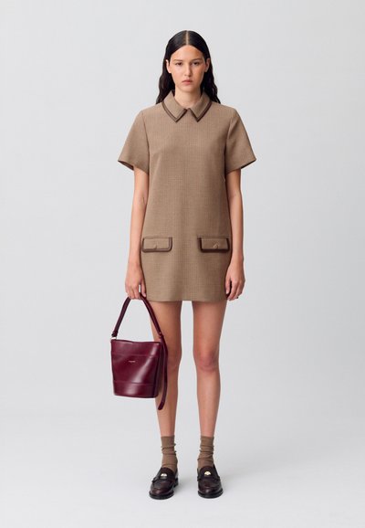 Giovane donna in un vestito a maniche corte beige a quadretti, che tiene in mano una borsa in pelle bordeaux, indossando calze marroni e mocassini scuri, in piedi su uno sfondo neutro.