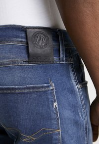 Blå jeans i denim med ett texturerat lädermärke i midjan som har en präglad logotyp och text. Jeansens slitna finish och gula sömnadsdetaljer är synliga.