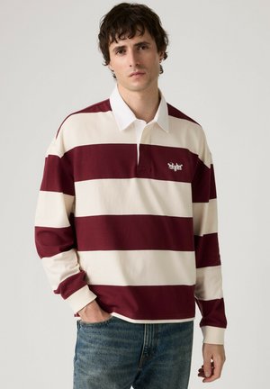 Mann trägt ein rugbyshirt mit breiten, burgunderroten und weißen Streifen, langen Ärmeln und weißem Kragen, dazu blaue Jeans, steht vor einem einfarbigen Hintergrund.