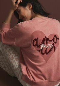 Sweatshirt roz dintr-un material moale, cu un grafic mare în formă de inimă și textul "amour" scris cu litere îndrăznețe și întunecate. Manșete și decolteu cu nervuri.