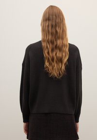 Schwarzer gerippter Pullover im übergroßen Design mit überschnittenen Schultern und einer strukturierten Oberfläche. Langes, gewelltes Haar ist sichtbar. Minimalistischer Hintergrund.