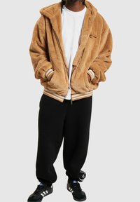 Brązowa kurtka z futerkiem z imitacji, z zamkiem z przodu, dużym kapturem i paskowanymi mankietami. Noszona na białej koszuli, w połączeniu z czarnymi dresami i butami.