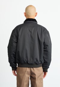 Jachetă bomber neagră cu talie și guler ribbed, având o textură netedă și o croială ușor supradimensionată. Purttată deasupra unei perechi de pantaloni bej.