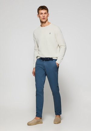 White long-sleeve shirt, blue chinos, and beige espadrilles. Shirt features a small logo; chinos have a smooth finish and side pockets.