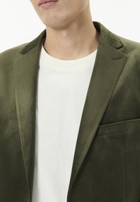 Olivově zelený semišový blazer s úzkým střihem, jednou kapsou a vyhrazenými klopami, nošený přes bílý hřejivý crewneck tričko s žebrováním.