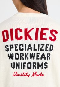 Pull crème avec texte tricoté en rouge et noir : "DICKIES UNIFORMES DE TRAVAIL SPÉCIALISÉS Fabriqué avec qualité." Encolure côtelée.