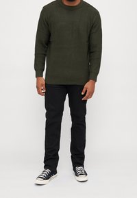 Man som står och bär en mörkgrön stickad tröja, svarta byxor och svartvita sneakers mot en neutral vit bakgrund.