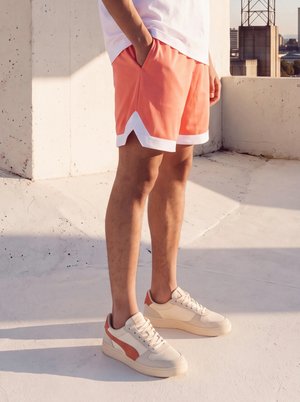 Une personne portant un short corail et des baskets blanches se tient sur une terrasse ensoleillée sur un toit, avec vue sur les immeubles de la ville.
