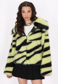 Faux fur jakke med neongrønne og sorte striber, der har lynlås fortil, blød tekstur og en stor krave. Sættes sammen med en sort plisseret nederdel.