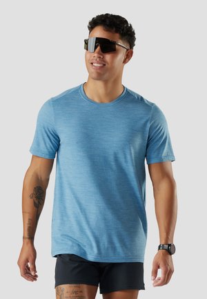 Man wearing a blue short-sleeve shirt, black shorts, large black sunglasses, and a black watch, smiling against a gray background.
