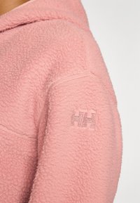 Nahaufnahme eines pinken Fleece-Hoodies, die eine weiche Textur und das gestickte 'HH'-Logo am Ärmel zeigt und das gemütliche Design hervorhebt.