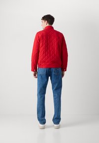 Rode gewatteerde jas met een kraag, knopen en een gladde textuur, gecombineerd met blauwe jeans en witte sneakers. Achteraanzicht wordt getoond.