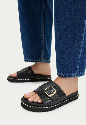 Fødder iført sorte slide-sandaler med guldbeslag og guldbeklædte kanter på sålerne, kombineret med blå denimjeans, der er afskåret over anklen.