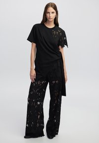 Top negro con detalles de encaje asimétricos en un lado, combinado con pantalones de encaje negros de corte holgado que presentan patrones florales y recortes.