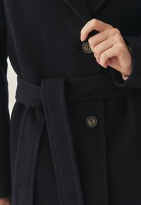 Marineblauer Wollmantel mit Taillengürtel, ausgestattet mit zwei Knöpfen, einem Reverskragen und einer weichen Textur, die klassische Designelemente hervorhebt.