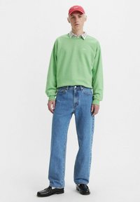 Sudadera verde con cuello redondo, combinada con jeans rectos de un azul claro. Una gorra roja y zapatos de vestir negros complementan el conjunto.