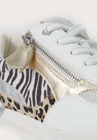 Baskets blanches avec des accents métalliques dorés, fermeture éclair latérale, panneaux imprimés zèbre et léopard, et lacets blancs sur fond blanc.