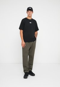 Sort t-shirt med korte ærmer og hvidt logo, olivengrønne bukser med knæpaneler og sorte sko. Modellen står foran en ensfarvet baggrund.