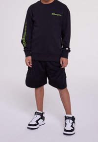 Black sweatshirt with green logo on chest and sleeve, paired with black mesh shorts. Footwear features a black and white design with straps.