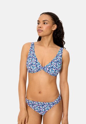 Mujer vistiendo un bikini azul con estampado floral, con un top con detalle de nudo en el frente y bragas a juego, de pie contra un fondo claro y liso.
