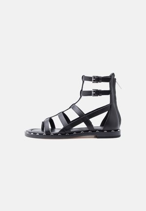 Sandale negre plate, stil gladiator, cu două curele la gleznă, catarame argintii, nituri de-a lungul marginii tălpii și închidere cu fermoar la spate.