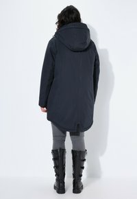 Dlouhá tmavě modrá bunda s kapucí, strukturovanými rukávy a mírně zakřiveným lemem. Nositá přes šedé kalhoty a černé vysoké boty.