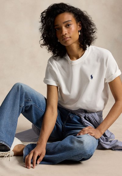 Młoda kobieta o kręconych włosach, ubrana w białą koszulkę z krótkim rękawem, niebieskie dżinsy i białe trampki, siedząca na podłodze w zrelaksowanej pozie.