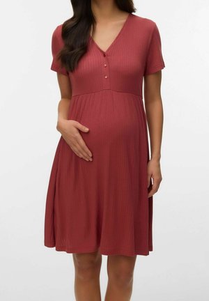 Zwangere vrouw die een rood geribd jurkje met korte mouwen en knopen draagt, haar buik met één hand vasthoudend.