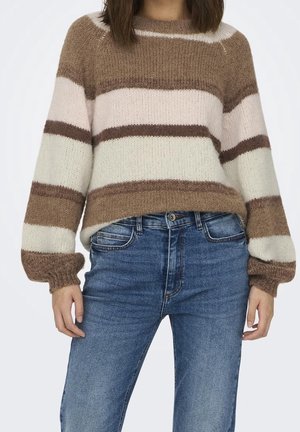 Personne portant un pull en laine rayé marron, beige et blanc à manches longues et un jean bleu, debout devant un fond uni.