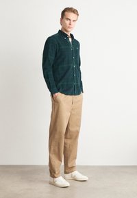 Camisa de flanela xadrez verde, com colarinho abotoado e mangas longas; combinada com calças bege de corte solto e ténis brancos.