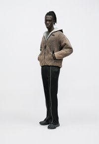 Mann trägt eine braune Fleecejacke mit Reißverschluss und Kapuze, schwarze Hose mit weißen Seitenstreifen und schwarze Sneakers, steht vor einem einfarbigen hellen Hintergrund.