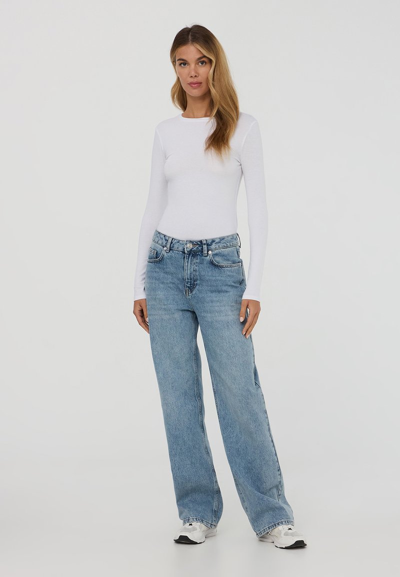 Top a maniche lunghe bianco aderente abbinato a jeans a gamba larga blu chiaro e sneakers bianche. Tessuto morbido con scollatura rotonda classica e design a cinque tasche.