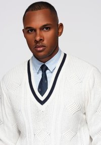 Maglione bianco lavorato a maglia con scollo a V profondo, con bordi blu navy. Sotto, una camicia colletto azzurra e una cravatta a motivo scuro.