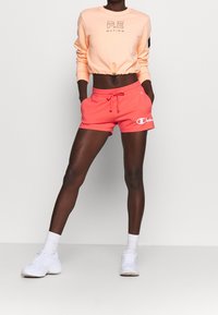 Korallfärgade shorts med dragsko i midjan och vitt Champion-logotyp, ihop med en ljusorange kort ärmad sweatshirt med svart P.E. Nation-logotyp.