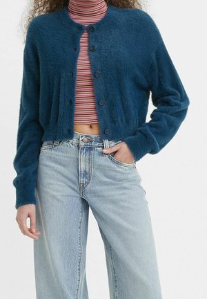 Femme portant un cardigan bouclé turquoise boutonné sur un col roulé rayé court, assorti à un jean taille haute bleu clair.