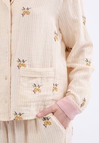 Conjunto de pijama de color amarillo claro con rayas verticales rosas, con bordado floral en amarillo y rosa. Incluye un bolsillo frontal y puños rosas.