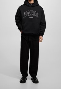 Sudadera de algodón negra con la palabra "PARIS" impresa en blanco, gran bolsillo frontal, combinada con pantalones de pierna ancha negros y zapatos negros.